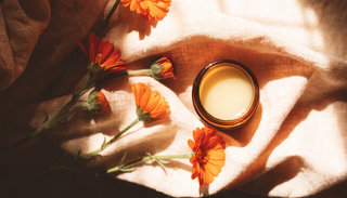 Fresh calendula flowers beside an open jar of golden balm on cream linen